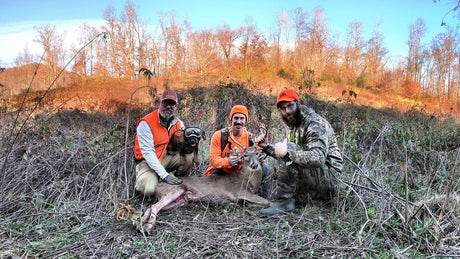 Three men in hunting gear posing with their deer and a dog wearing Rex Specs dog goggles.